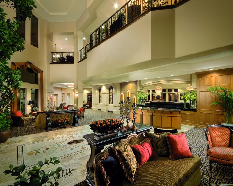 A spacious and elegantly decorated living room area in a senior living facility with high ceilings, large indoor plants, ornate furniture including sofas and chairs with patterned cushions, a decorative table with candles, and a reception desk in the background with floral arrangements and warm lighting.