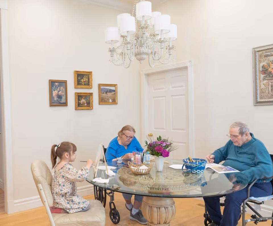 A young girl and two elderly individuals sitting around a glass dining table in a well-lit room. The elderly woman is painting, and the elderly man is looking at a book or magazine. The table has art supplies and a vase with purple flowers. The room has light-colored walls, framed paintings, a white door, and a chandelier hanging from the ceiling.