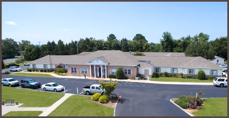 A single-story senior living facility building with a light brown exterior and a gray shingled roof, surrounded by a paved parking lot with several parked vehicles. The building has a central entrance with white columns and a small landscaped area with bushes and trees in front. The sky is clear and blue.
