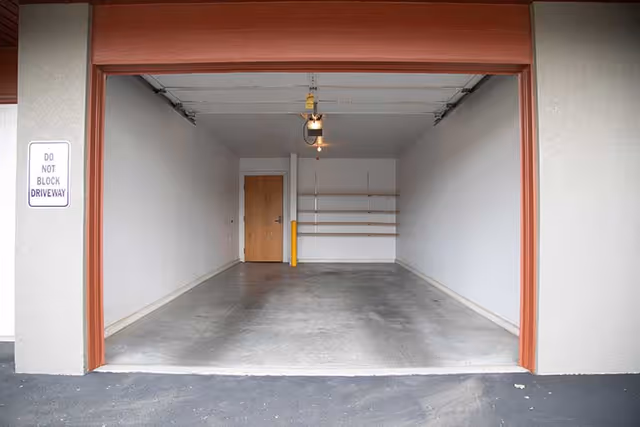 Empty single-car garage with concrete floor, wall-mounted shelves, ceiling light, and a closed wooden door at the back.