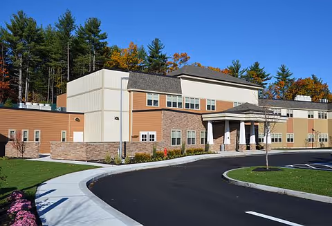 Exterior view of a modern assisted living facility building with a curved driveway, landscaped grass areas, and trees in the background under a clear blue sky.