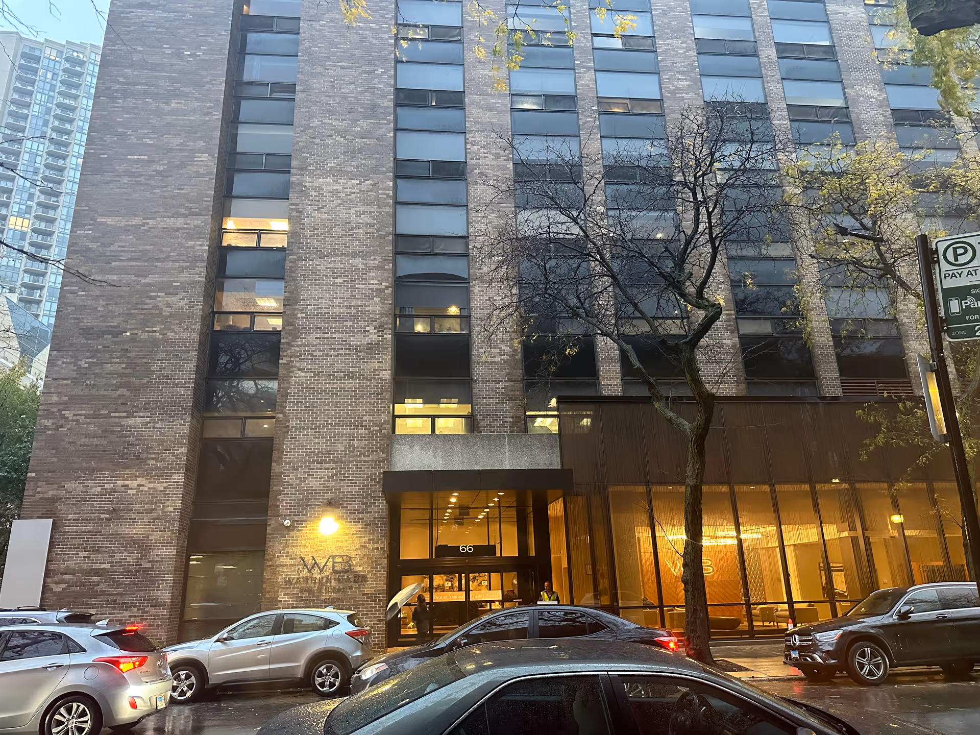 Exterior view of a multi-story brick building with large windows and a glass entrance. Several cars are parked along the street in front of the building. A tree with sparse leaves stands on the sidewalk near the entrance. The building has the letters 'WB' on the wall near the entrance, and the address number 66 is visible above the door.