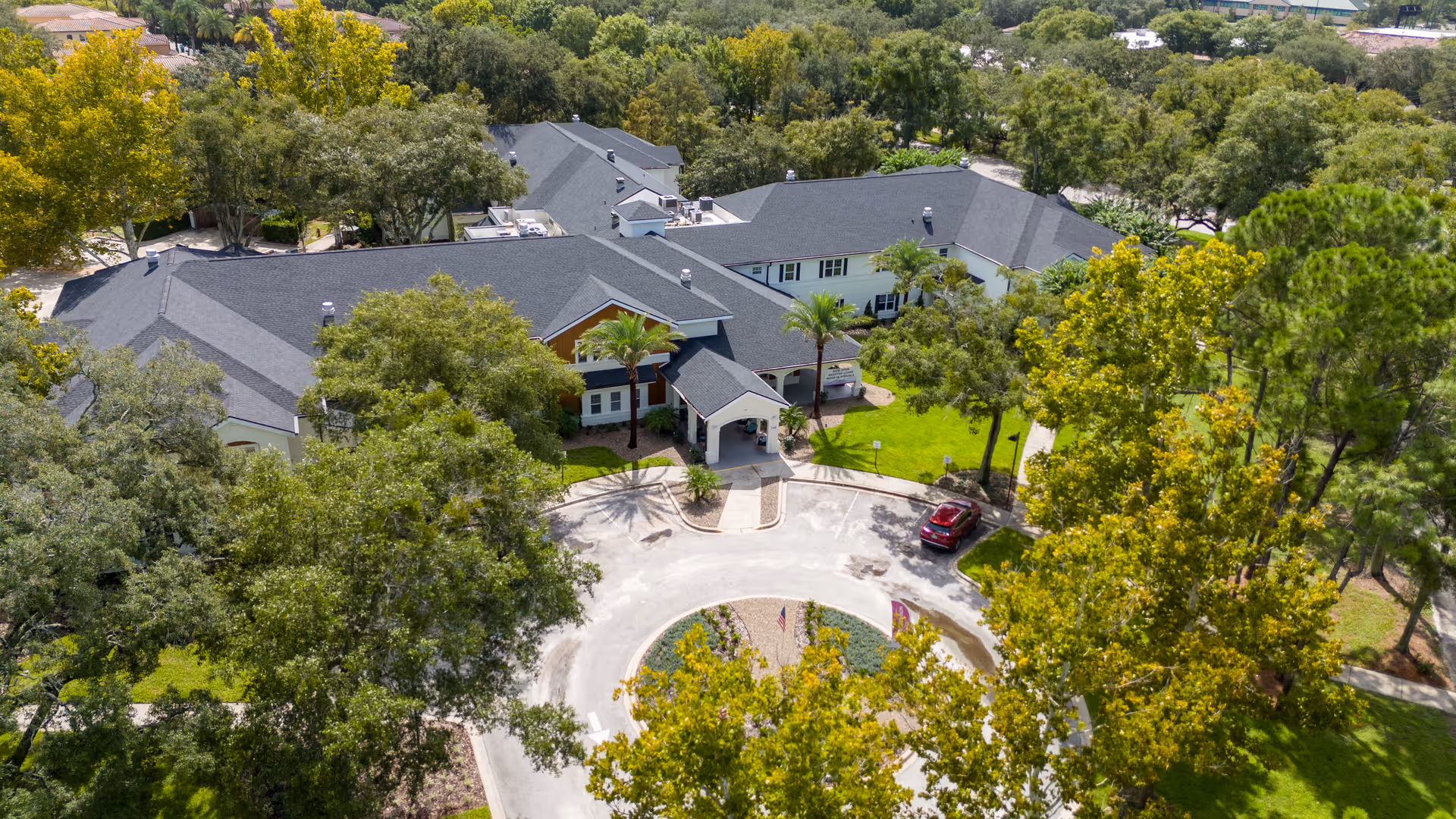 Aerial view of Ambiance at Maitland senior living facility showing a large building with a dark gray roof surrounded by green trees and landscaping. There is a circular driveway with a red car parked near the entrance, and the area is lush with greenery and well-maintained grounds.