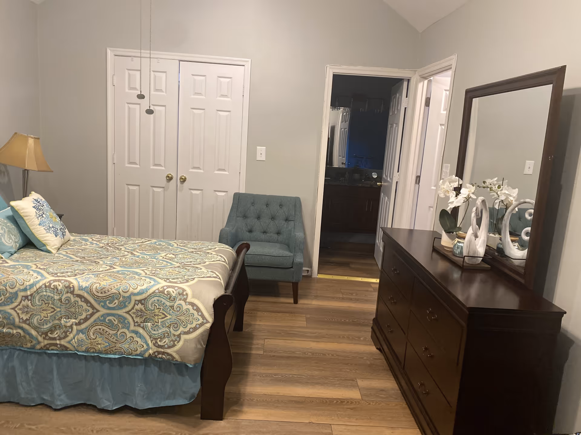 A bedroom with a bed covered in a patterned blue and beige bedspread, a blue upholstered chair, a wooden dresser with a large mirror, and decorative items including white flowers and sculptures. There are two white double doors and an open door leading to a bathroom.