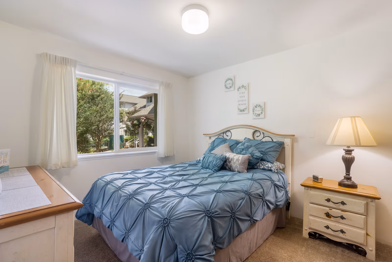 Bright bedroom with a blue quilted bed, a nightstand and lamp, a dresser, and a window showing trees outside.