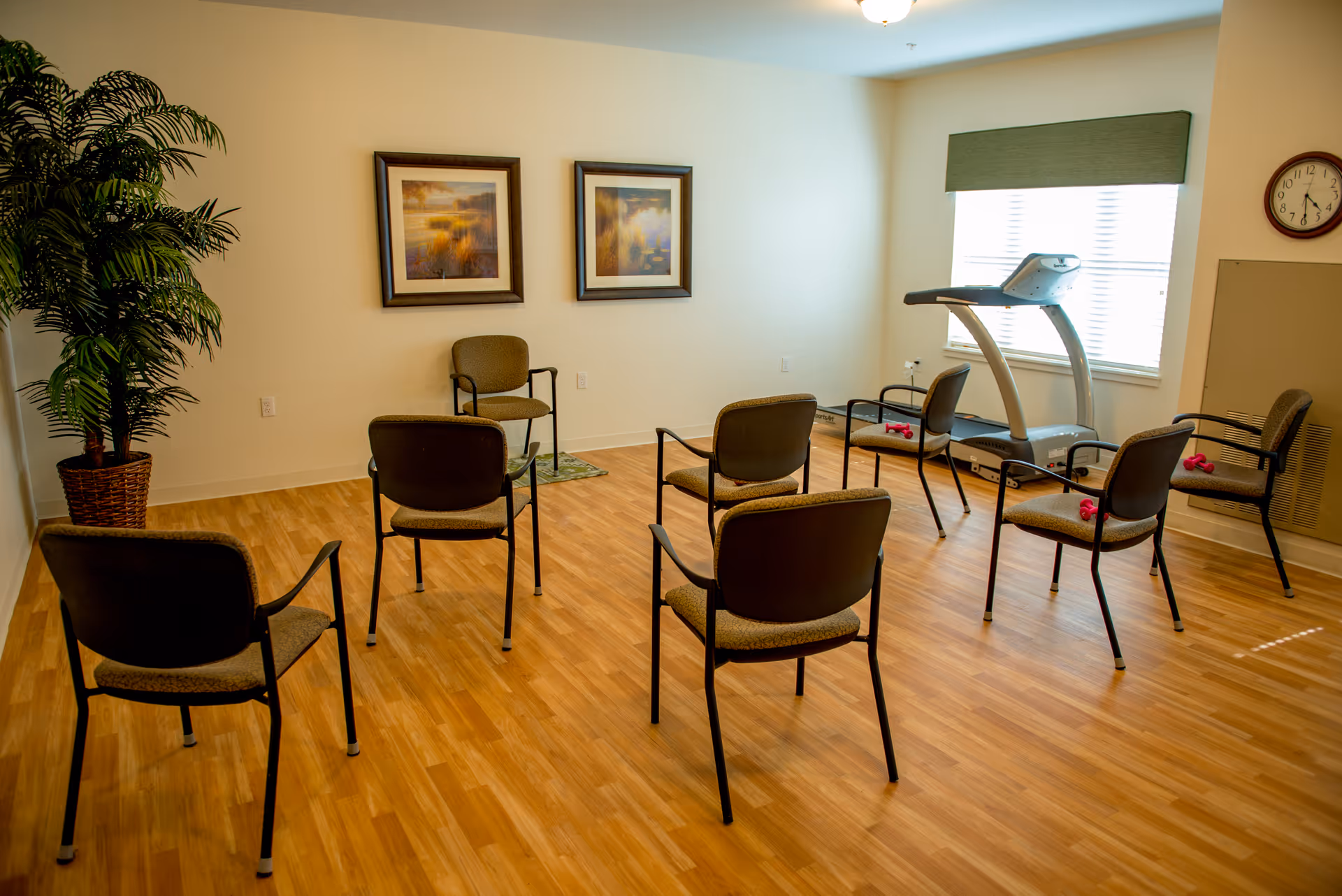 Small activity room with several arranged chairs, a treadmill by the window, wall art, a potted plant and a light wood floor.