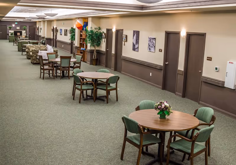 Interior hallway of a senior living facility with round tables and green cushioned chairs arranged along the carpeted corridor. The walls are beige with brown trim, decorated with framed pictures and wall sconces. There are several doors along the hallway and a potted plant near a bookshelf with colorful beach balls on top.
