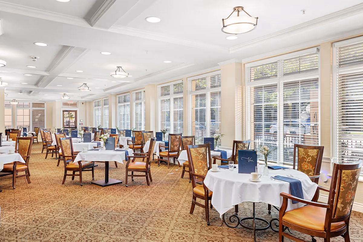 Bright dining room with round tables covered in white tablecloths and wooden chairs arranged along large windows.