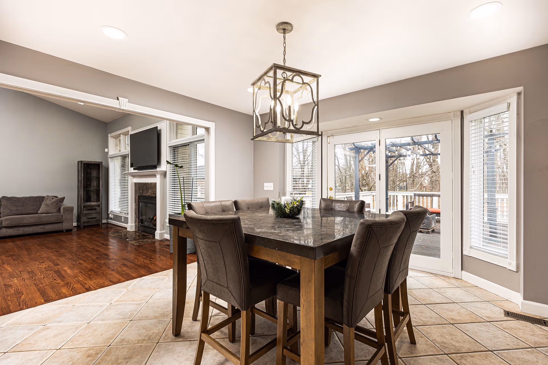 A modern dining area with a square marble-top table surrounded by six cushioned chairs. The room has beige tiled flooring and gray walls. A decorative hanging light fixture is above the table. Large windows and glass doors provide a view of an outdoor deck with trees in the background. Adjacent to the dining area is a living room with wooden flooring, a gray couch, a fireplace with a mounted TV above it, and a tall shelving unit.
