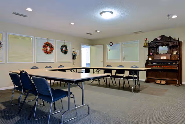 A meeting or activity room with tables arranged in a U-shape surrounded by blue chairs. The room has beige walls, three windows with closed blinds, and two decorative wreaths hanging on the wall. There is a wooden antique organ or cabinet against the far wall with small framed items on it. The ceiling has recessed lighting and a central light fixture.