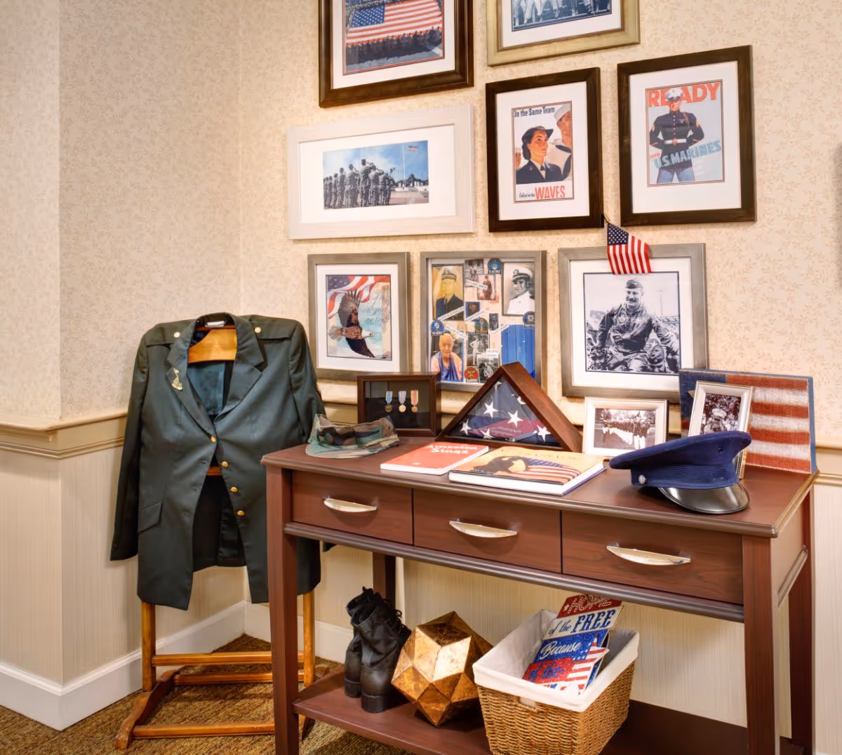 A corner display in a room featuring a military uniform jacket on a wooden stand, a wooden table with military boots, a folded American flag in a display case, framed photographs and posters related to the military on the wall, a military hat on the table, and a basket with patriotic-themed items underneath.