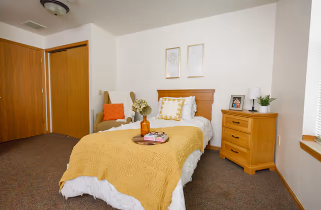 A cozy bedroom with a single bed covered in white bedding and a mustard yellow knitted blanket. The bed has decorative pillows and a wooden headboard. Next to the bed is a wooden nightstand with a lamp, a framed photo, and a small plant. There is a beige armchair with an orange cushion in the corner near a closet with wooden sliding doors. The room has carpeted flooring and two framed floral artworks hanging above the bed.