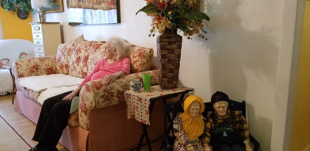 An elderly woman with white hair wearing a pink top and black pants is sitting on a floral-patterned couch in a cozy living room. Next to the couch is a small table with a crocheted tablecloth, a large woven vase with artificial flowers, and two cups. On the floor beside the table are two decorative dolls dressed in old-fashioned clothing. The room has light-colored walls and wooden flooring.