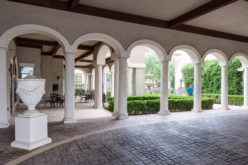 Covered outdoor walkway with white arches and columns, leading to a patio area with tables and chairs. There is a large decorative white urn on a pedestal and neatly trimmed green bushes. A person in a blue shirt is walking near the bushes.