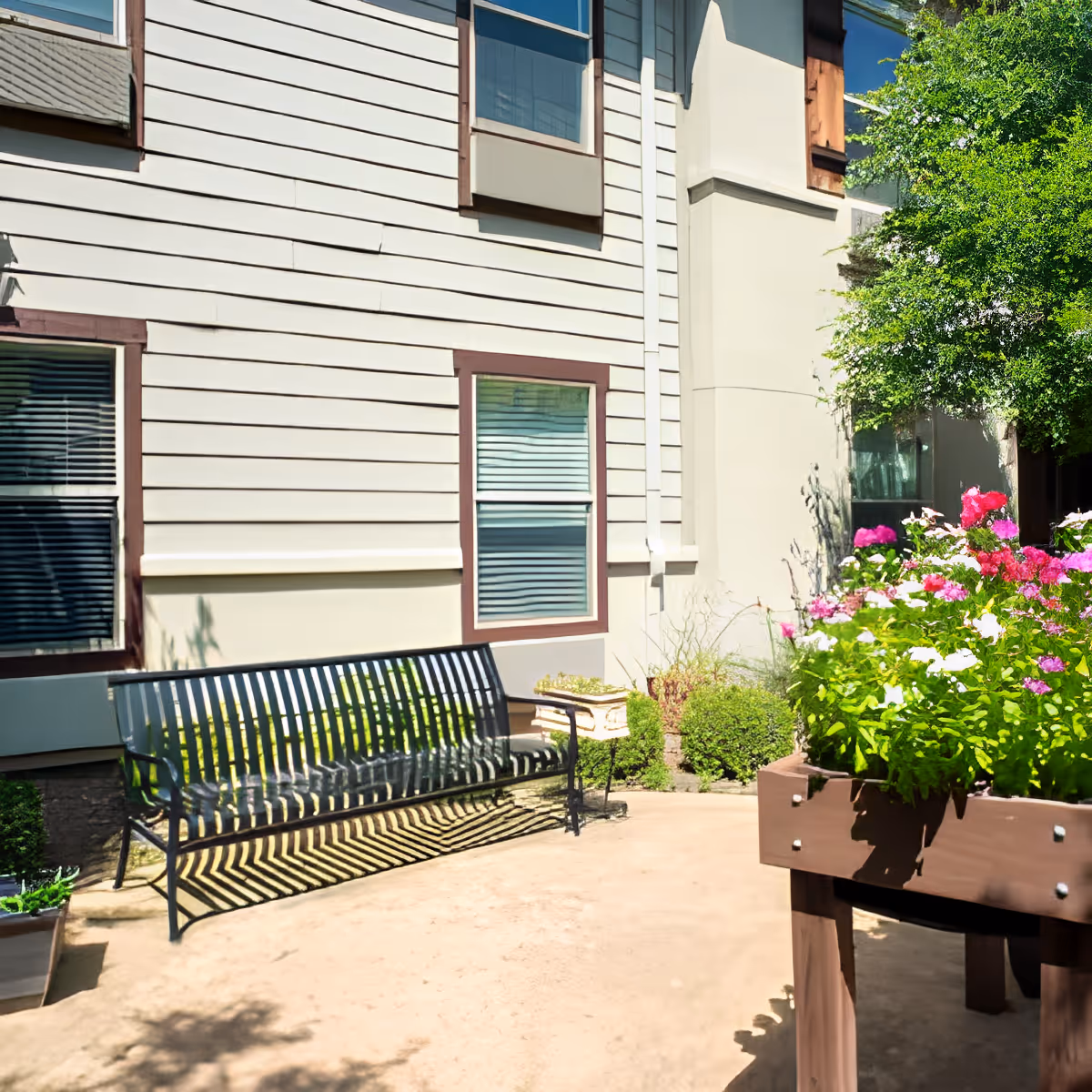 Outdoor patio area at Discovery Village At Southlake featuring a black metal bench, a raised wooden planter box filled with colorful flowers, and a building exterior with windows and beige siding.