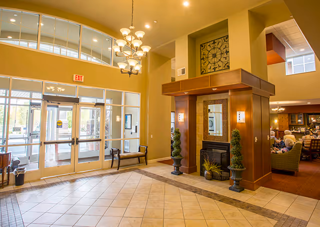 Bright senior living facility lobby with glass entrance doors, chandelier, seating, and a decorative fireplace area.