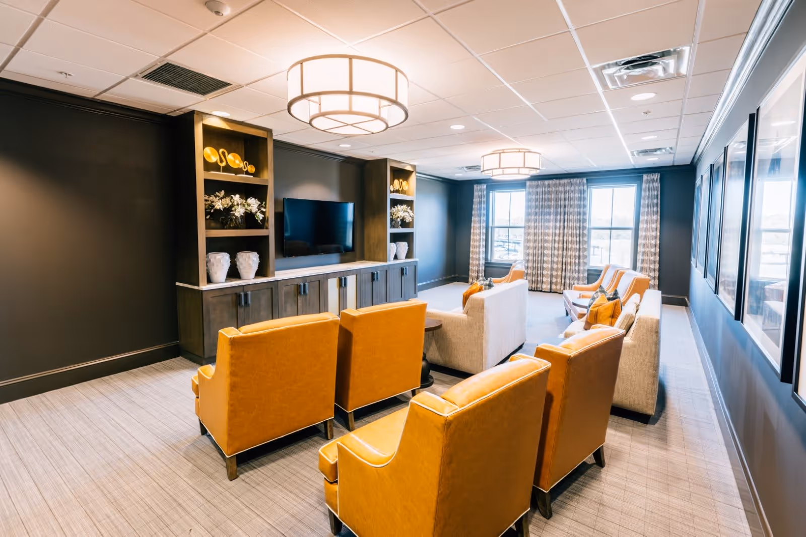A modern living room with mustard yellow armchairs and beige sofas arranged facing a wall-mounted flat screen TV. The room features built-in wooden shelves with decorative items, large windows with patterned curtains, and two round ceiling lights.