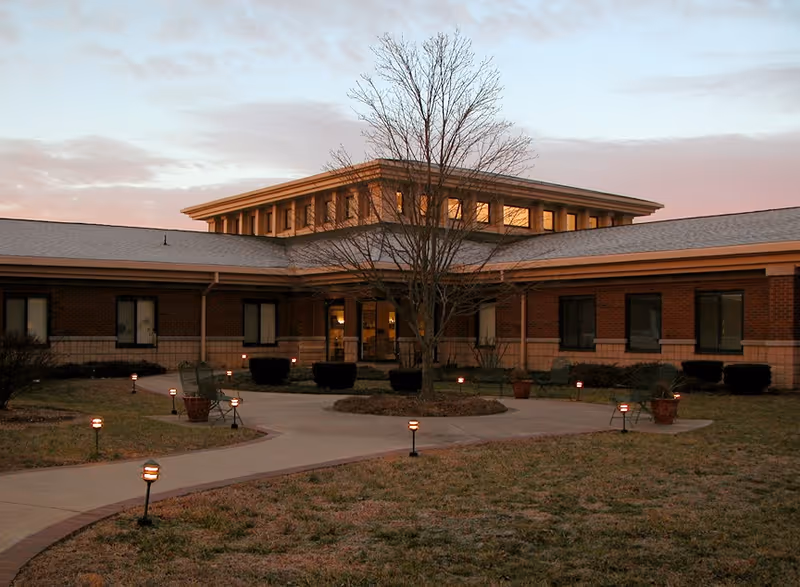 Front entrance of a single-story brick senior living building with a paved walkway, low landscape lights, and a leafless tree at dusk.