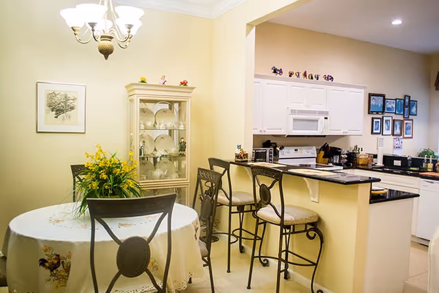 Interior view of a dining area and kitchen in a senior living facility. The dining area features a round table with a floral tablecloth and a centerpiece of yellow flowers, surrounded by metal chairs. Adjacent to the dining area is a kitchen with a breakfast bar, three high chairs, white cabinets, a microwave, stove, and various kitchen appliances. The walls are painted light yellow, and there is a glass cabinet displaying plates and decorative items.