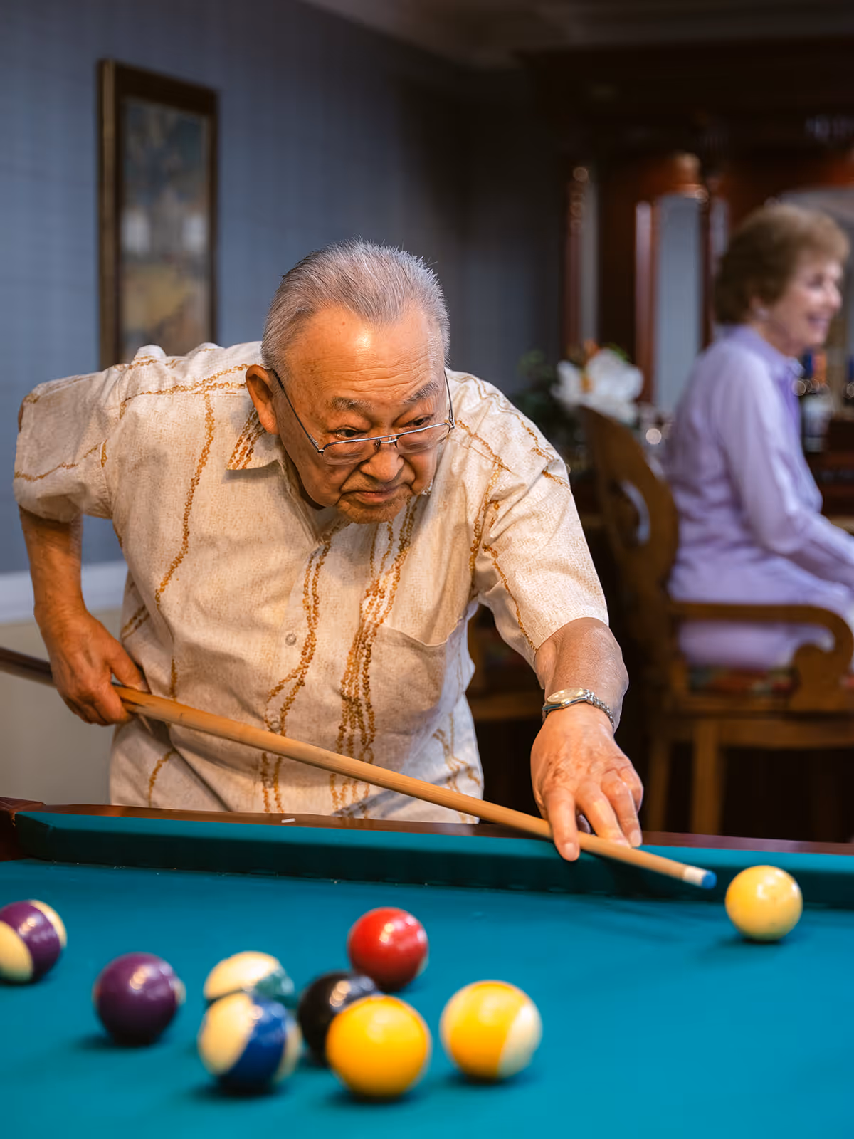 An elderly man wearing glasses and a beige patterned shirt is playing pool, aiming to hit the cue ball with a pool cue. In the background, an elderly woman in a lavender outfit is sitting on a wooden chair, smiling. The setting appears to be an indoor recreational area.
