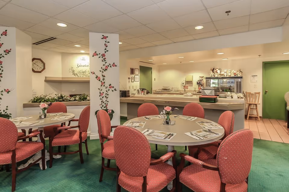 A cozy dining area with round tables set with placemats and small flower vases. The chairs are upholstered in red fabric with a subtle pattern. The room has green carpet flooring and white walls decorated with floral vine designs. In the background, there is a counter area with kitchen equipment and a sign that reads 'The Garden Cafe'.