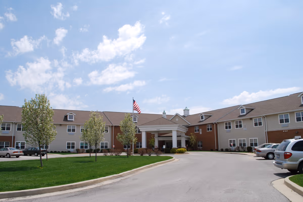 Exterior view of a two-story senior living facility building with a covered entrance, several windows, and an American flag on a flagpole in front. There are small trees and green grass along the driveway, with several parked cars visible on the right side.