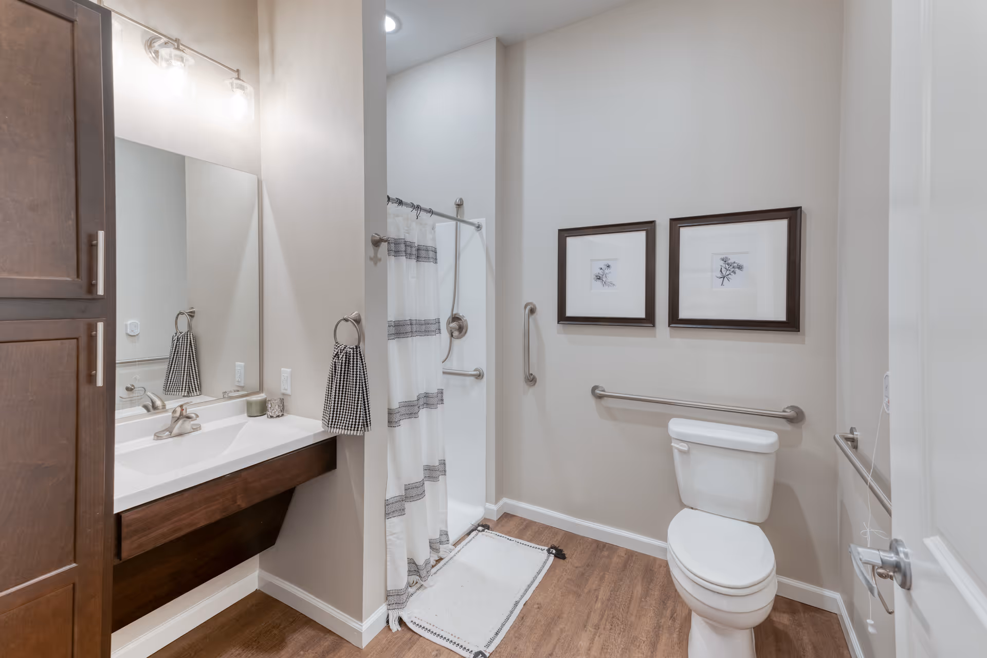 A clean and accessible bathroom with a white toilet, grab bars on the walls, a shower with a white curtain featuring gray stripes, a wooden vanity with a white sink, a large mirror, and two framed botanical prints on the wall.