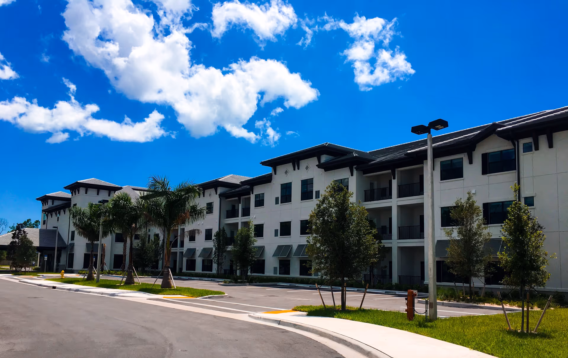 Exterior view of a modern three-story senior living facility building with balconies, palm trees, and a clear blue sky with scattered clouds.