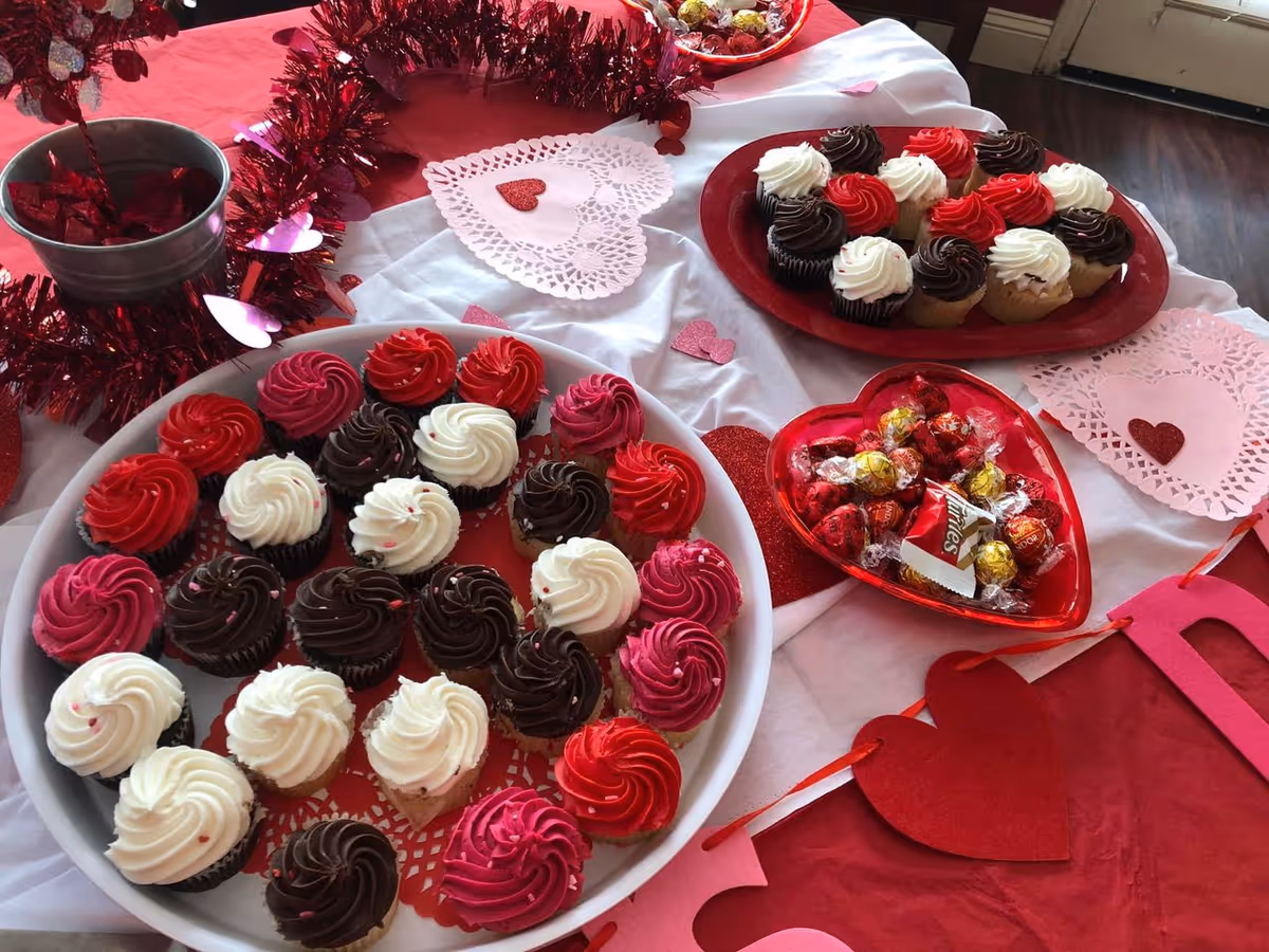 A festive Valentine's Day dessert table with trays of red, pink, white, and chocolate-frosted cupcakes and heart-shaped candies.