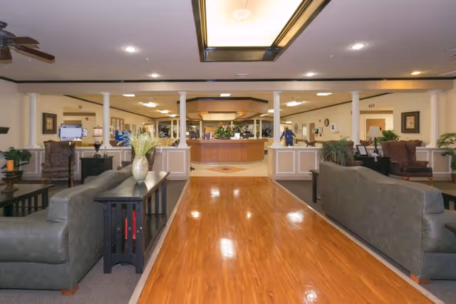 Interior view of a healthcare facility lobby with a polished wooden floor leading to a reception desk. The area is furnished with two gray sofas facing each other, side tables with decorative plants, and armchairs. The ceiling has recessed lighting and a ceiling fan, with columns separating the seating area from the reception.