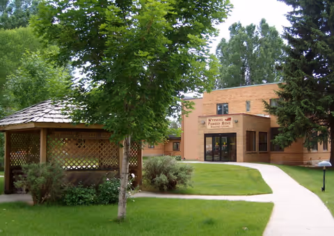 Exterior view of Wyoming Pioneer Home, a brick building with a sign above the entrance. In the foreground, there is a green lawn with a paved walkway and a wooden gazebo surrounded by trees and shrubs.