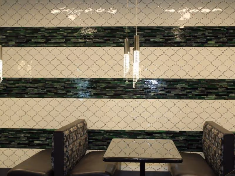 A dining booth with two cushioned seats facing each other and a square table in between. The wall behind has a decorative tile pattern with alternating horizontal bands of cream and dark green tiles. Two modern pendant lights hang above the table.