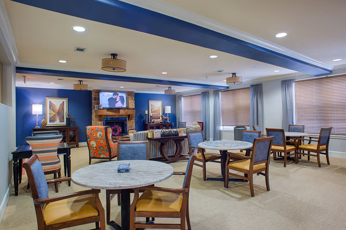 A well-lit common area with multiple round tables and chairs arranged for seating. The room features a blue accent wall with a stone fireplace and a mounted TV above it. There are framed artworks on the walls, large windows with blinds and curtains, and ceiling lights providing illumination.
