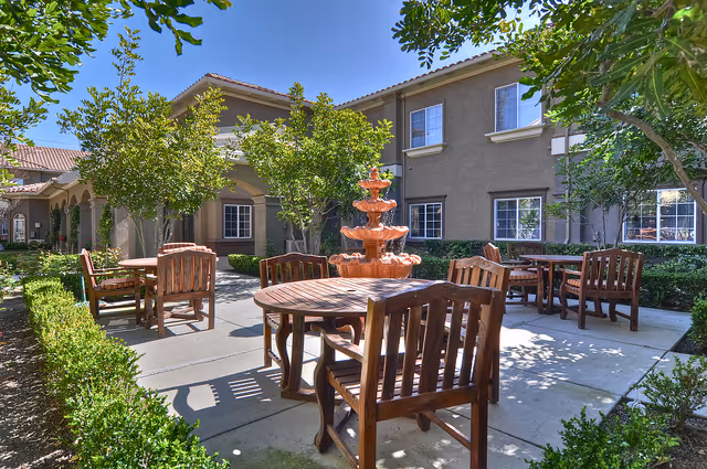 Sunny outdoor courtyard with wooden tables and chairs arranged around a central tiered fountain in front of a two-story building.