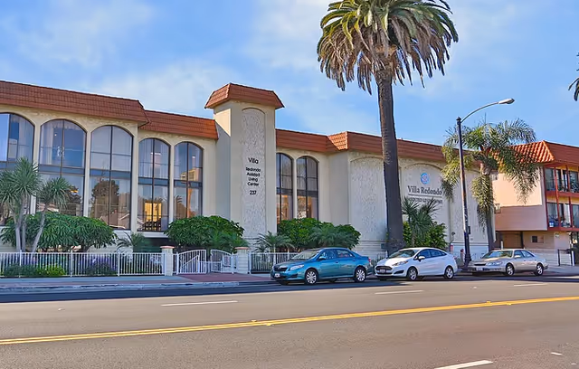 Front exterior of the Villa Redondo building with large arched windows, palm trees, and parked cars along the street.