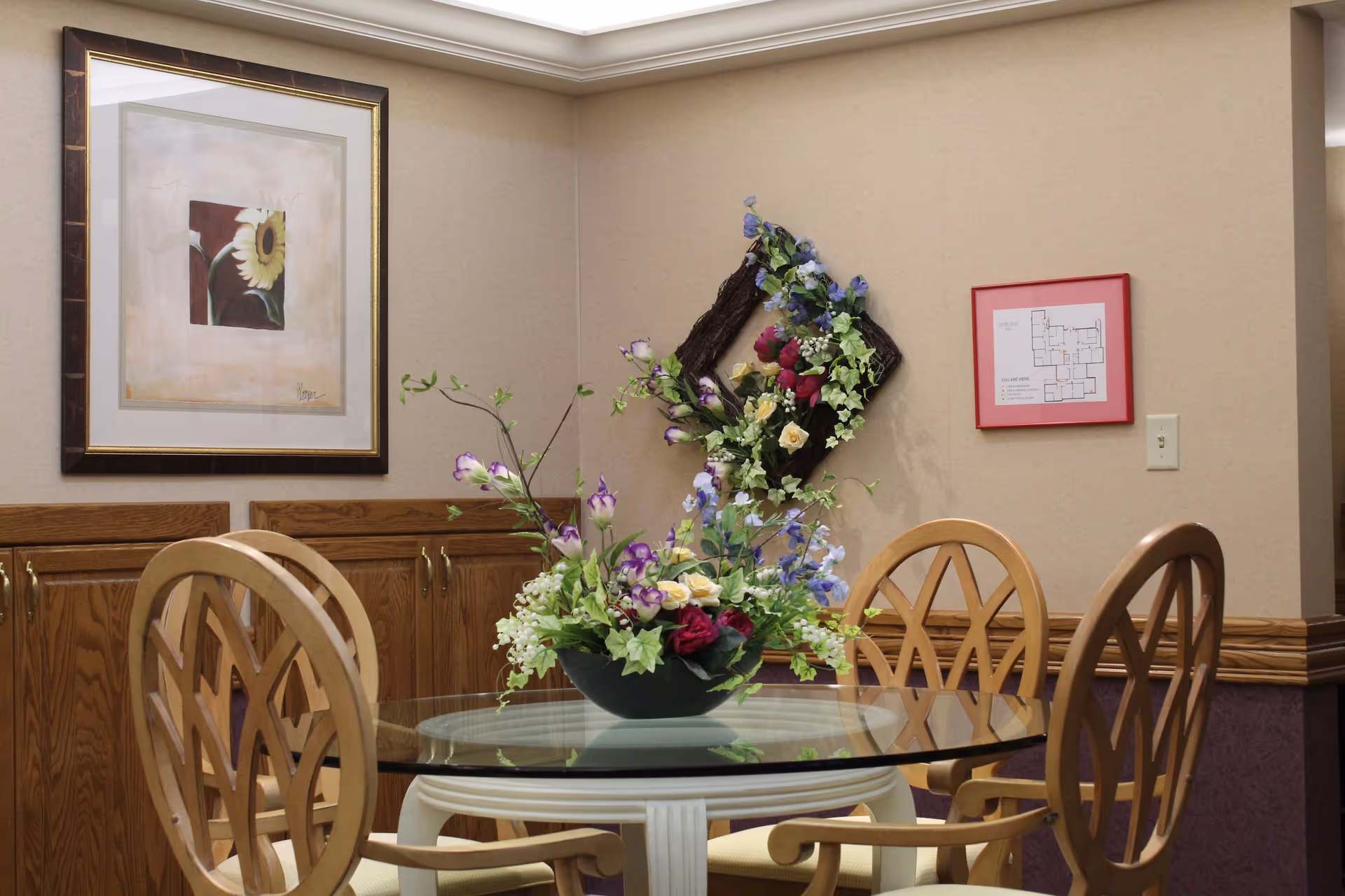 A small dining area with a round glass table surrounded by four wooden chairs with decorative backs. On the table is a floral arrangement with various colorful flowers. The walls are beige with wooden paneling on the lower half. There is a framed picture of a sunflower on the left wall and a floral wreath decoration on the back wall. A framed floor plan is also visible on the back wall.