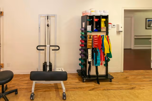 A small exercise area with a weight machine, a rack holding colorful dumbbells and resistance bands, and a wooden floor. The background shows a hallway with framed pictures on the wall.