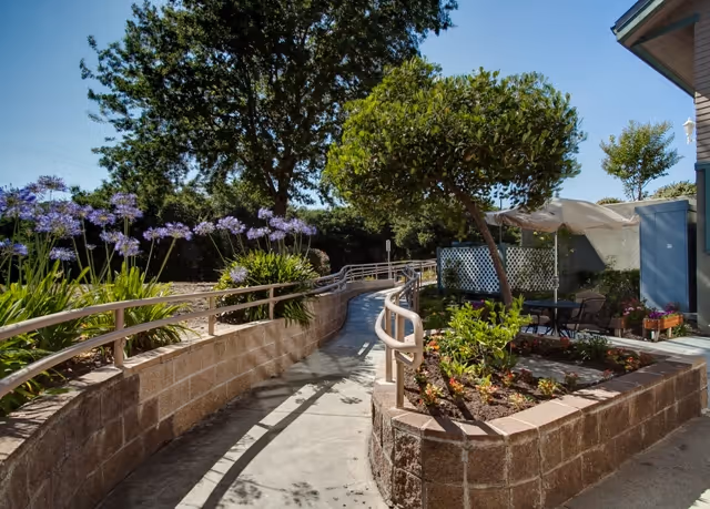 Outdoor garden area with a concrete pathway bordered by low brick walls and metal handrails, surrounded by plants and flowers. There is a small seating area with a table and chairs under a canopy, and trees providing shade in the background under a clear blue sky.