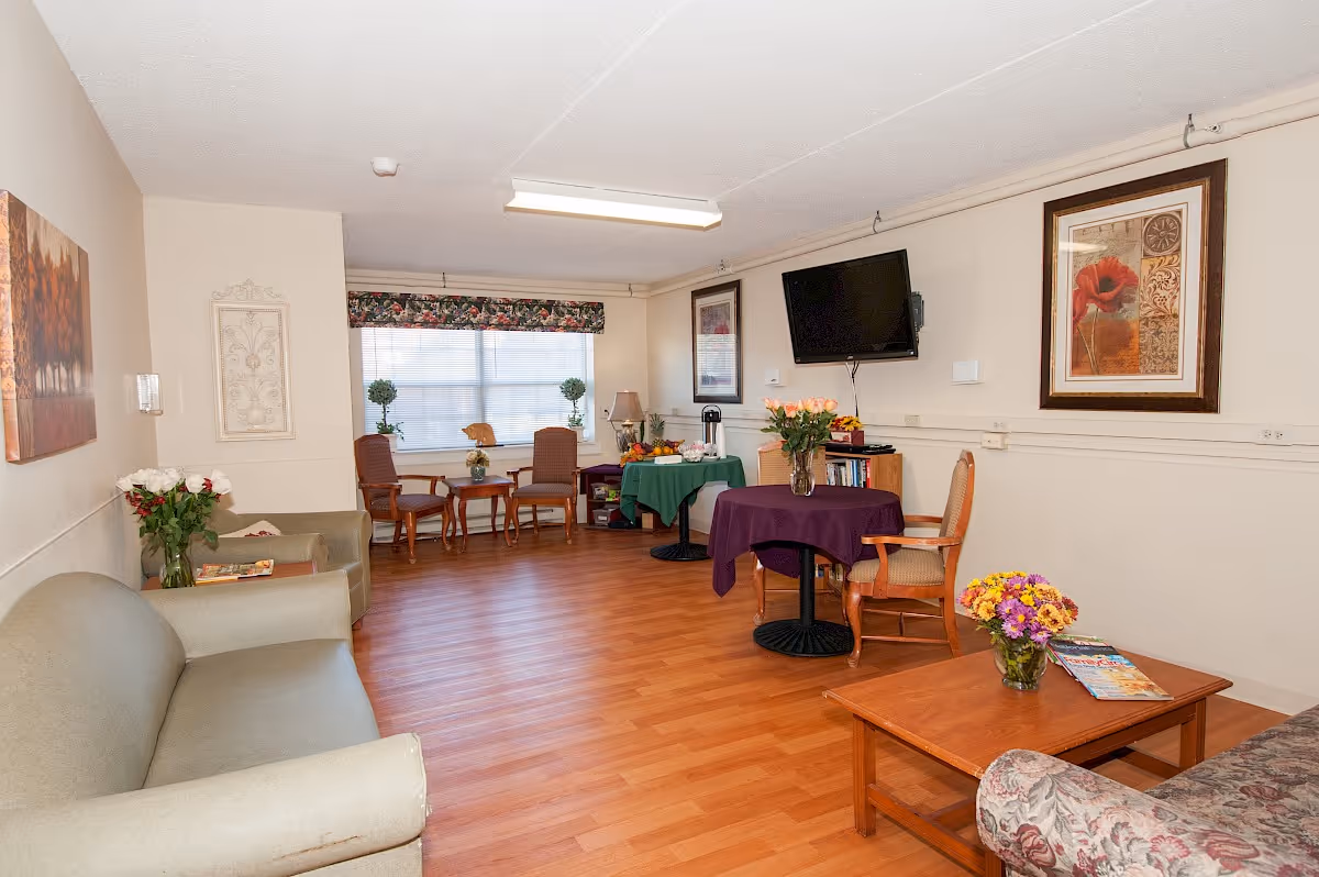 Bright communal living room with sofas, chairs, small tables, a wall-mounted TV, and floral arrangements.