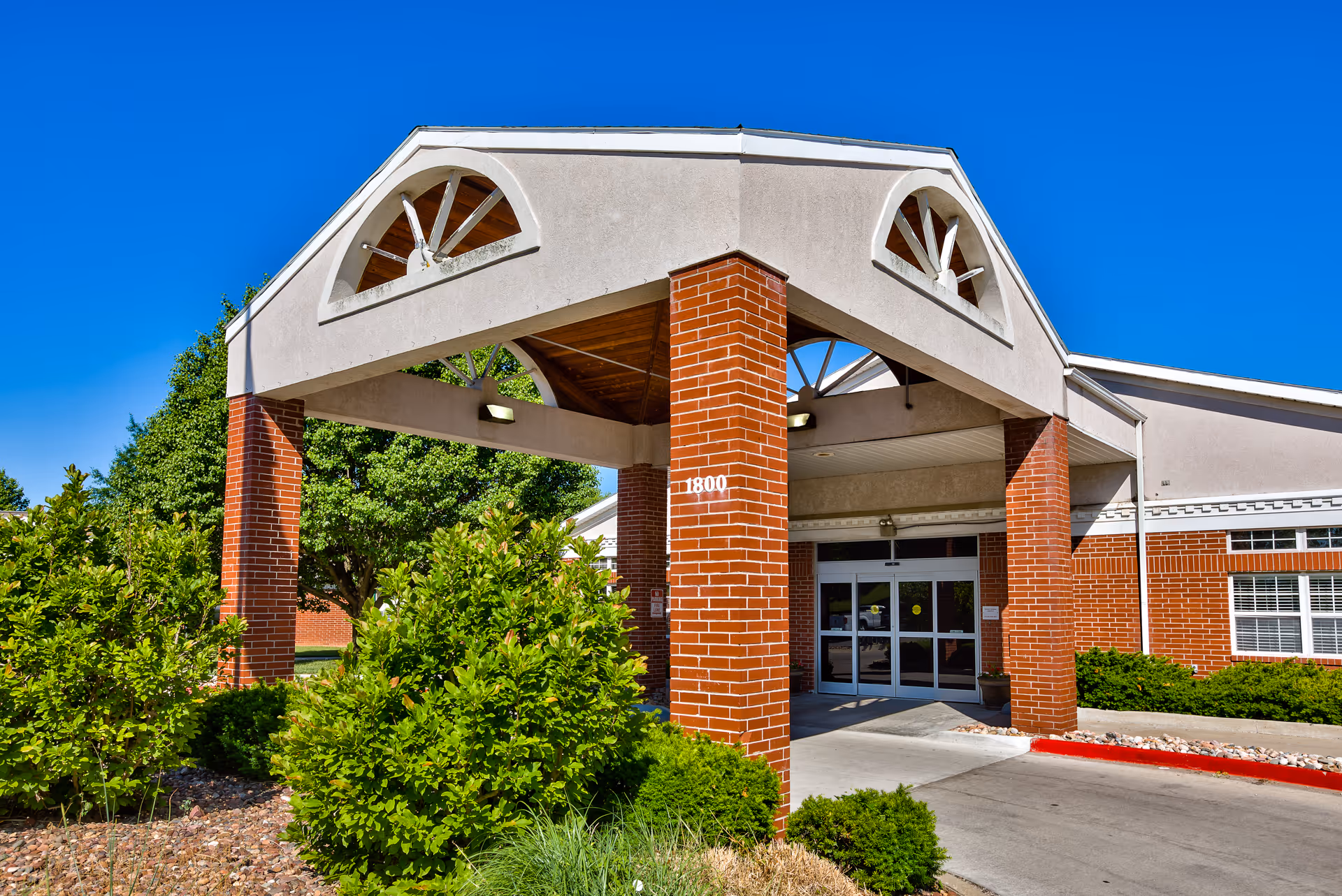 Covered entrance with brick columns and glass double doors at the front of a senior living facility, surrounded by shrubs under a clear blue sky.