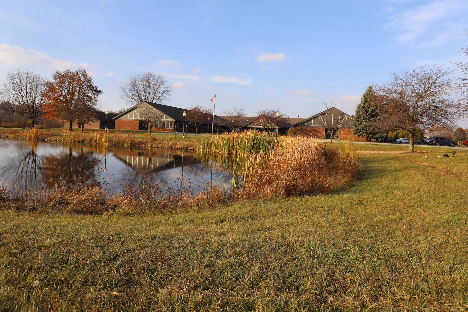 A scenic outdoor view of a senior living facility with a pond in the foreground reflecting the building and surrounding trees. The building has a brick exterior with triangular roof sections. Trees with autumn foliage and a grassy area surround the pond under a partly cloudy sky.