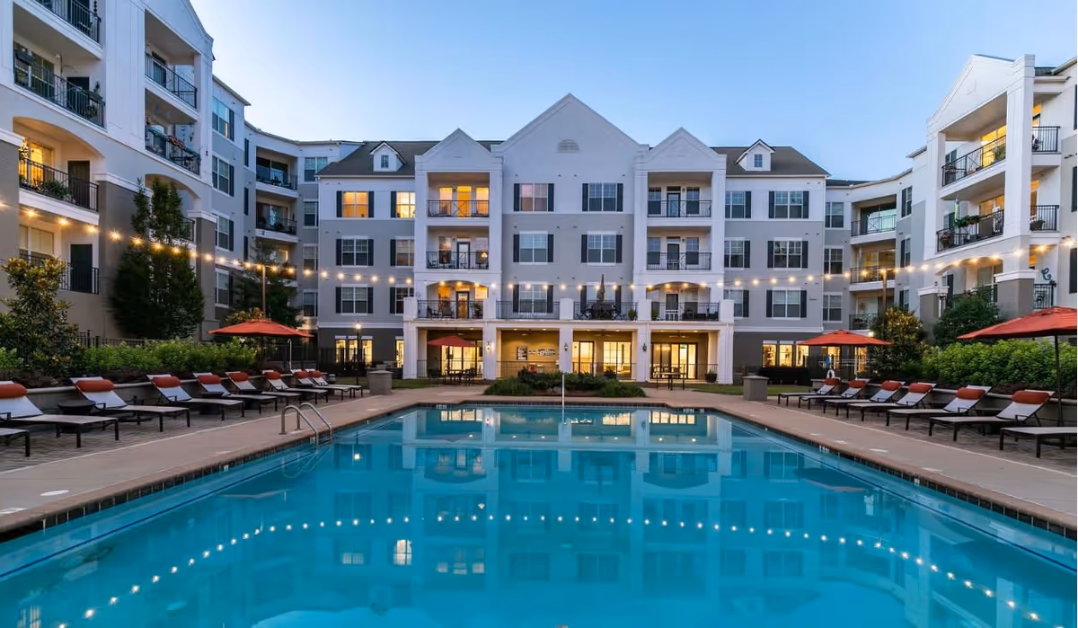 Outdoor courtyard with a central swimming pool surrounded by lounge chairs and a multi-story residential building lit at dusk.
