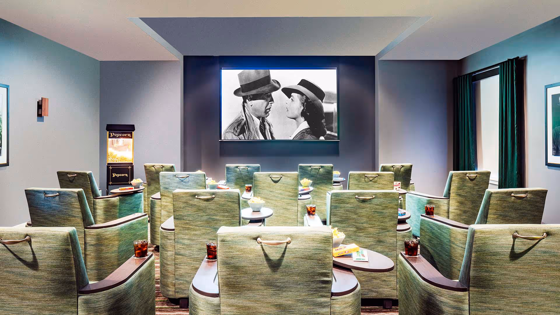 Small screening room with rows of green upholstered recliner chairs, small tables with drinks and snacks, a popcorn machine, and a black-and-white movie on a screen.