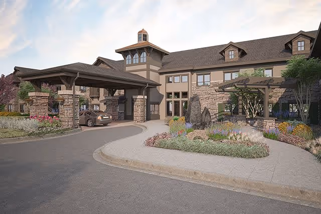 Exterior view of a senior living facility named The Peaks at Old Laramie Trail, featuring a large covered entrance with stone pillars, a circular driveway, landscaped garden beds with flowers and shrubs, and a multi-story building with multiple windows and dormers under a partly cloudy sky.