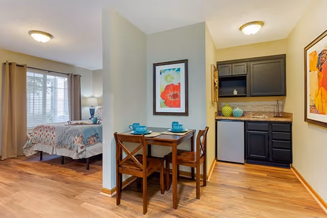 A cozy studio apartment interior featuring a small dining table with two wooden chairs set with blue plates and cups. To the right is a compact kitchenette with black cabinets, a small refrigerator, and decorative items on the counter. In the background, there is a bed with floral bedding near a window with beige curtains, allowing natural light to fill the room. The walls are painted light beige, and the floor is wood laminate.