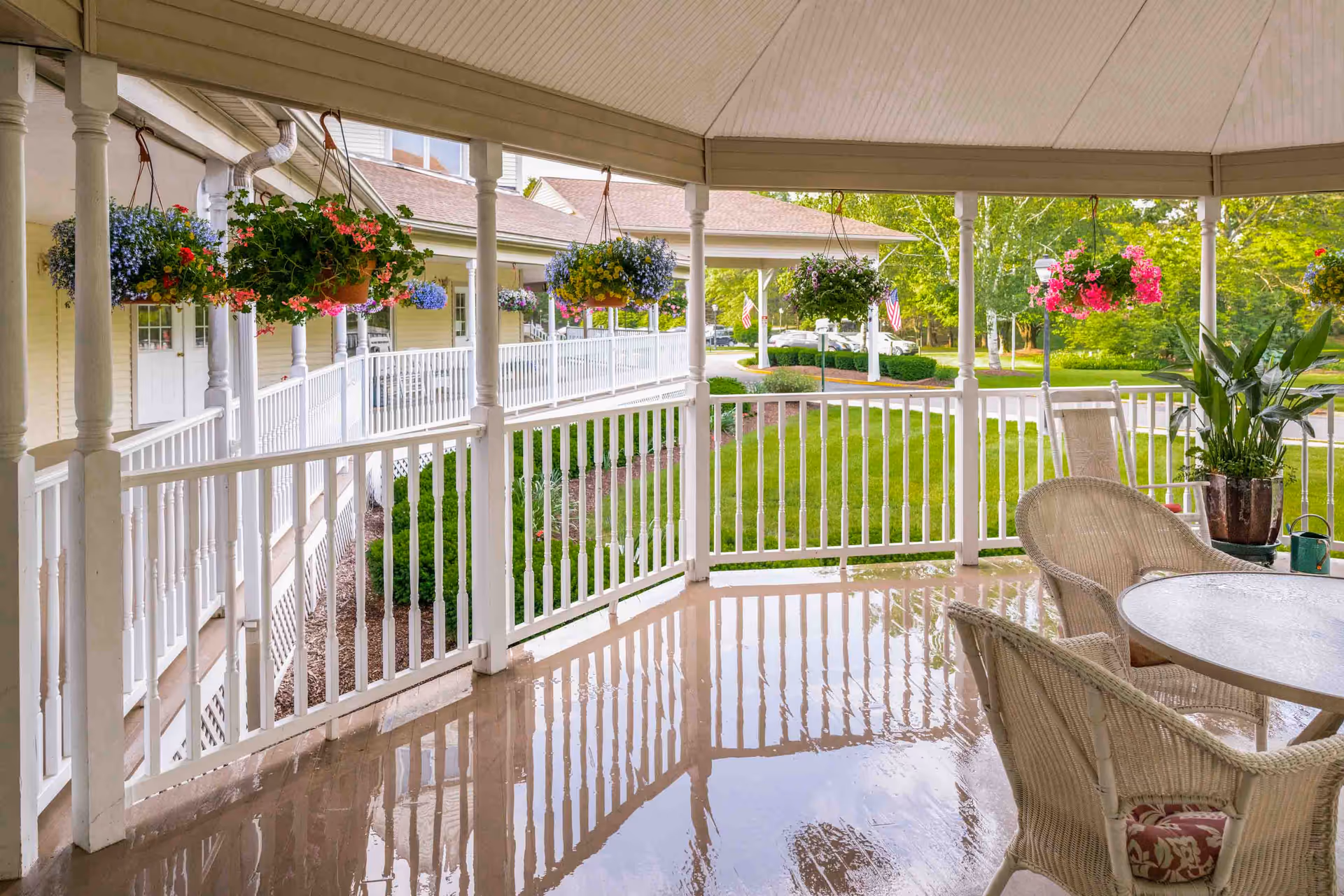 Covered porch with white railing, hanging flower baskets, and wicker chairs and table overlooking a green lawn.