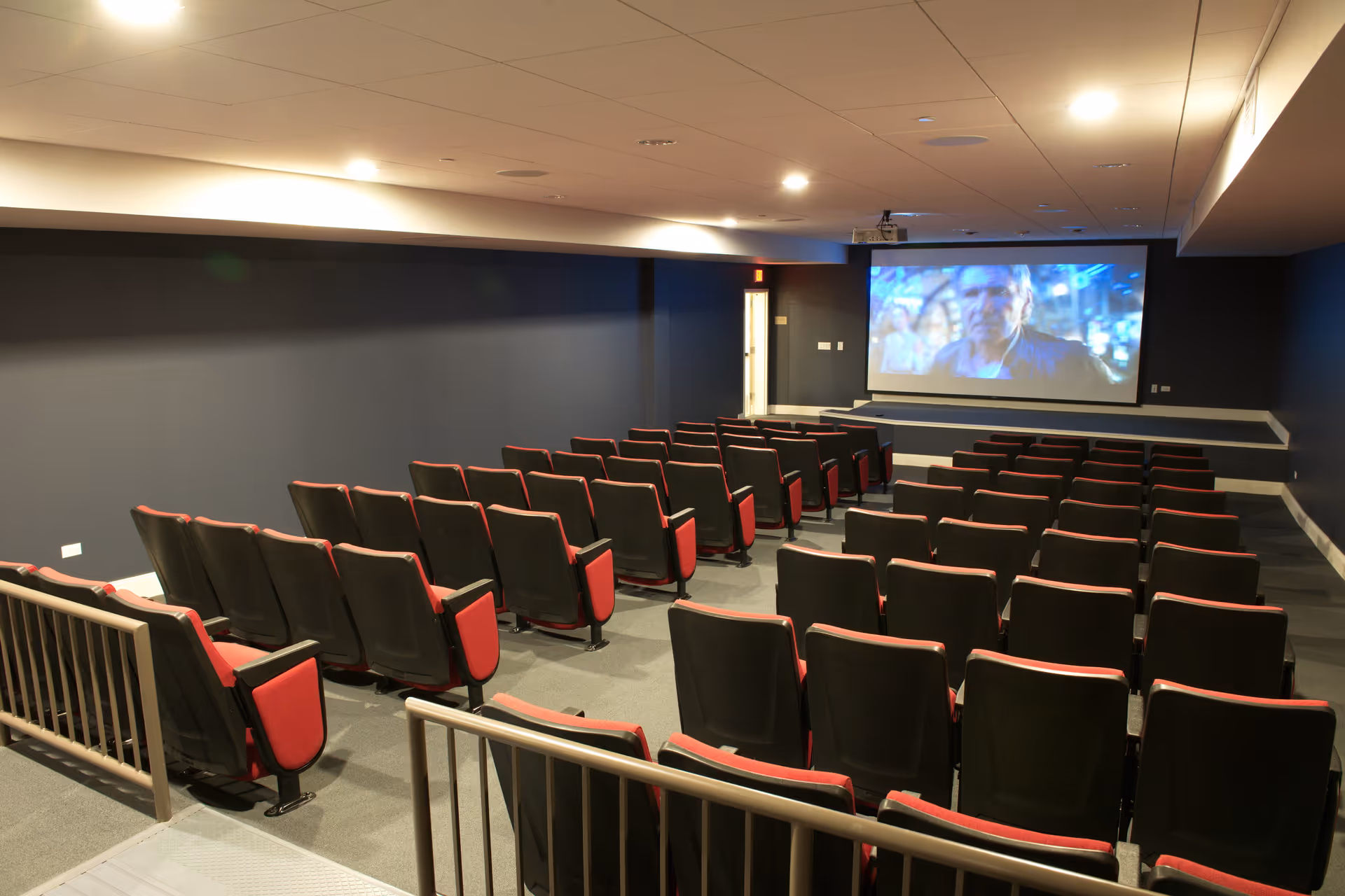 A small theater room with rows of black and red cushioned seats facing a large screen displaying a movie scene. The room has dark walls and ceiling lights, with a railing visible in the foreground.