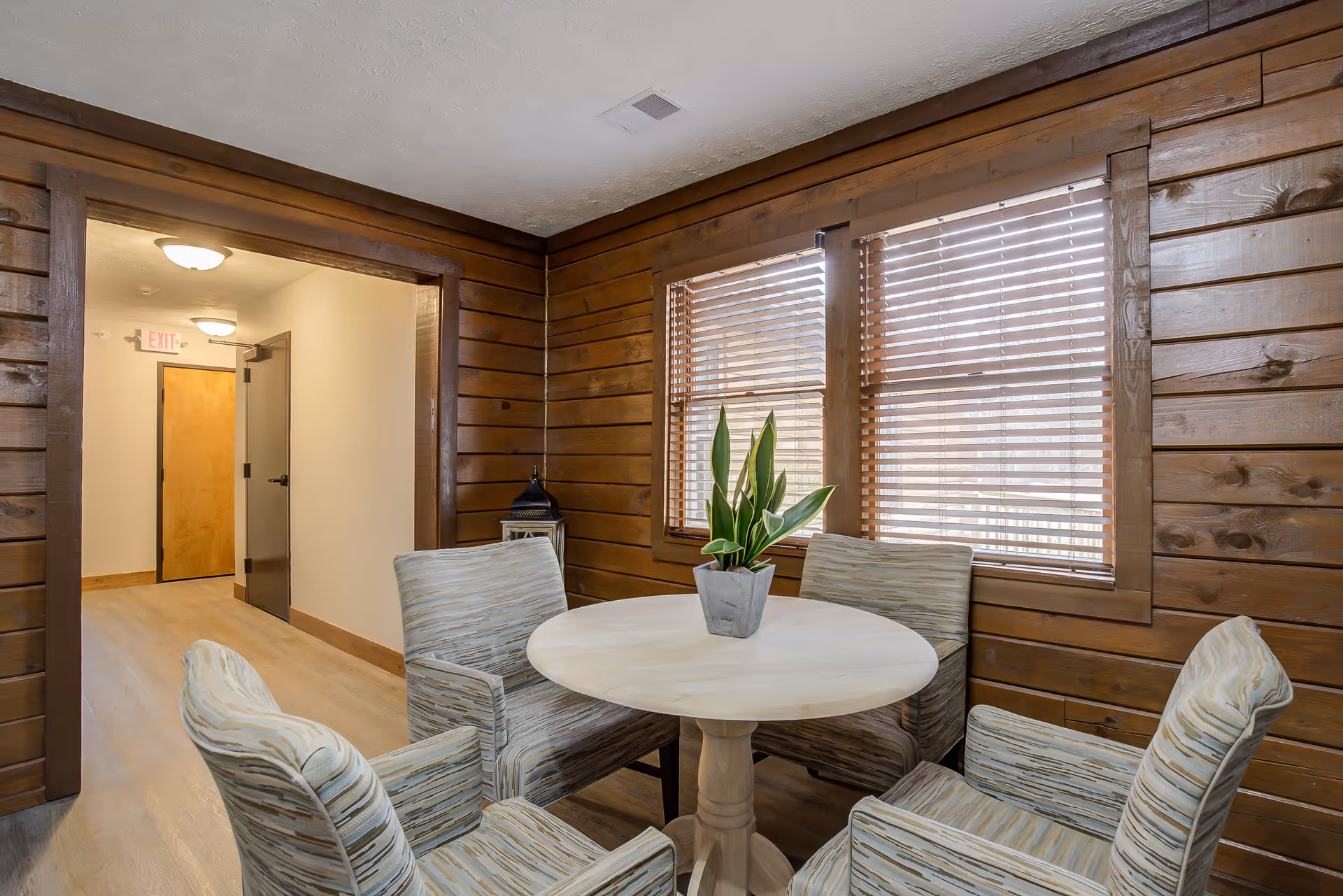 A cozy interior room with wood-paneled walls and two windows with wooden blinds. In the center is a round white table with a potted plant on top, surrounded by four cushioned armchairs with striped upholstery. A hallway with light-colored walls and wooden doors is visible through an open doorway.