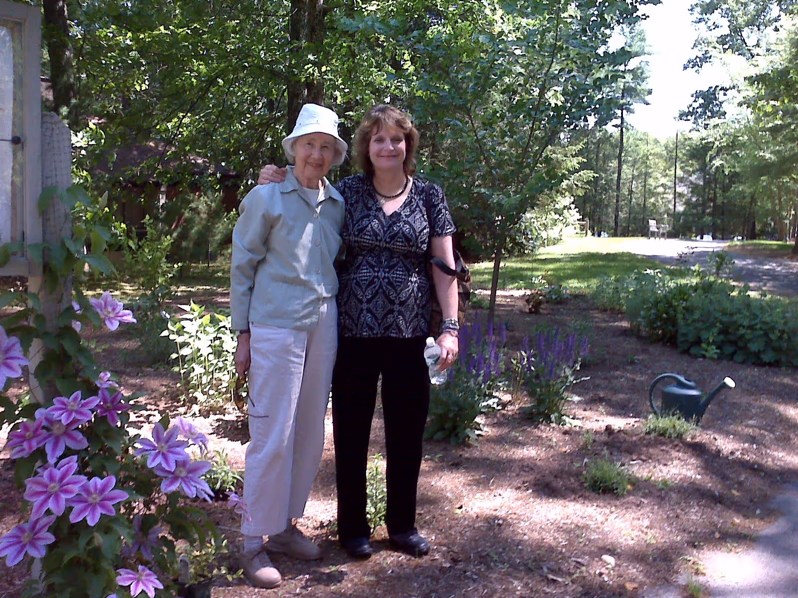 Two women standing outdoors in a garden area with trees and flowering plants around them. One woman is older, wearing a light-colored jacket, pants, and a white hat. The other woman is younger, wearing a patterned blouse and black pants, holding a water bottle. A green watering can is visible on the ground nearby.