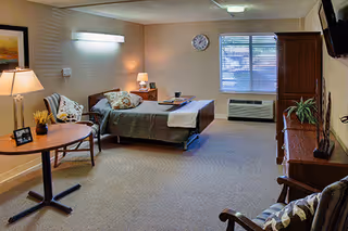 A furnished senior living suite featuring a bed, couch, desk, lamps, and a window.